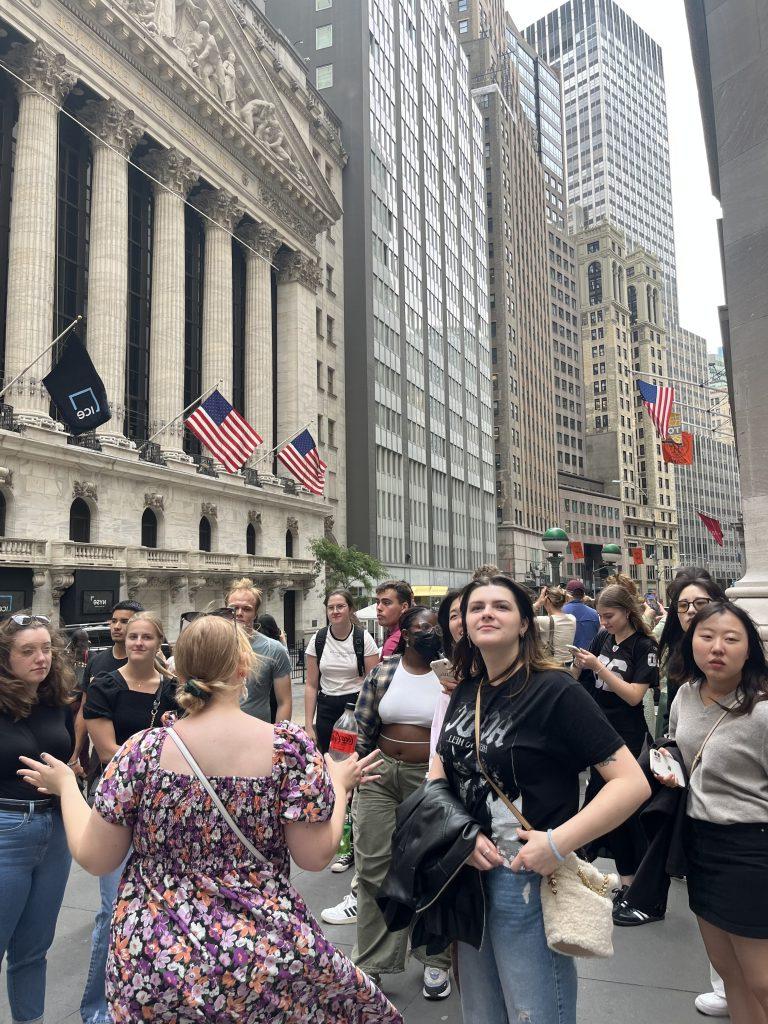 Group of students from different cultures and countries standing with their advisor on Wall Street who explains the history of the area