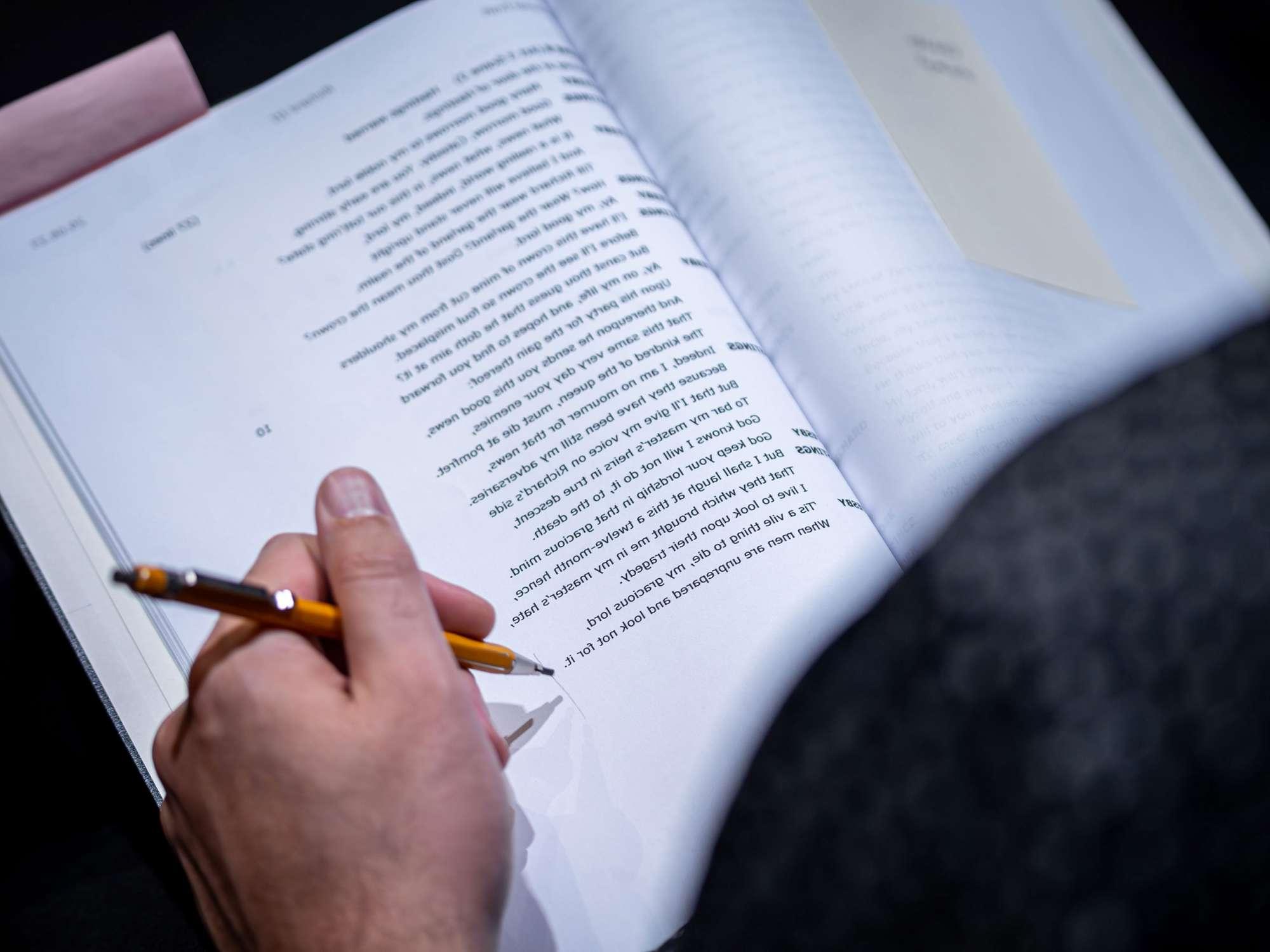 A hand with a pencil, follow along on a page from a Shakespeare play script.