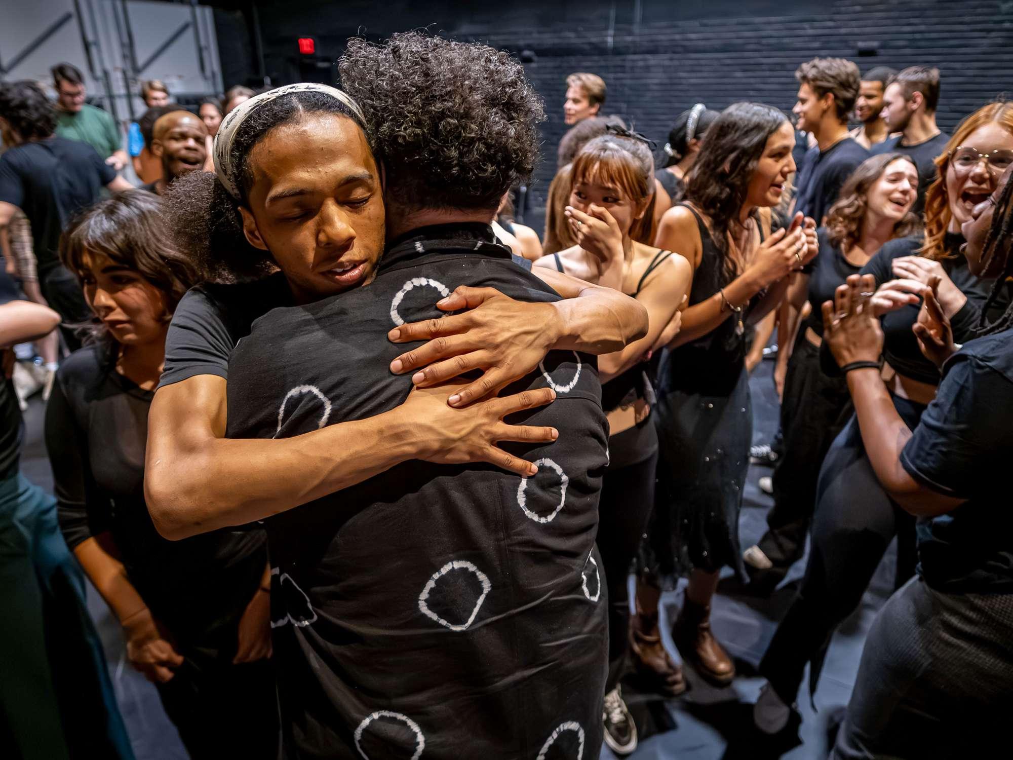 A student actor hugs the director while other student actors gather in the background.