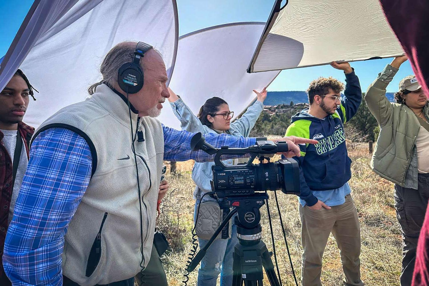 A professor standing by a video camera and wearing headphones instructs four students holding photo reflectors over their heads.