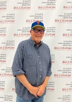 A man standing in front of a backdrop 与 writing. He has glasses, a blue hat and is wearing a button down dress shirt 与 jeans.