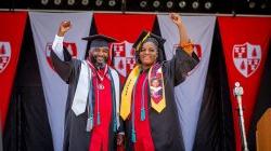 Father and daughter graduates, Lavone and Tiiera Broxton stand together with arms raised in their regaliaon Commencement stage with university banners behind them