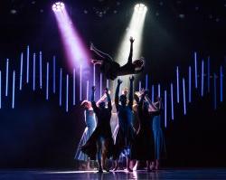 Group of dancers tossing another dancer into the air under spotlights